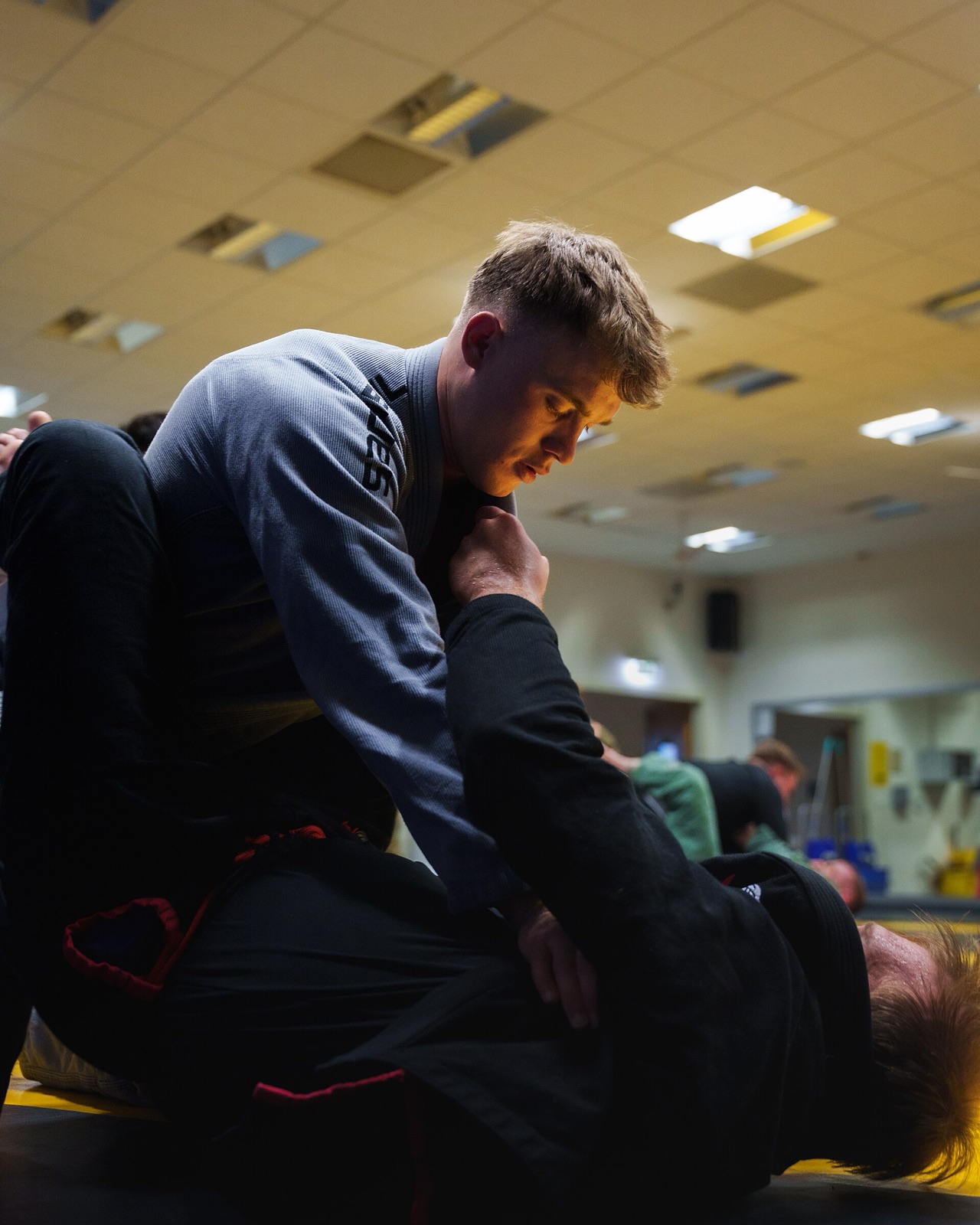 A grappler in a grey gi works from inside his partner's closed guard in a busy gym.