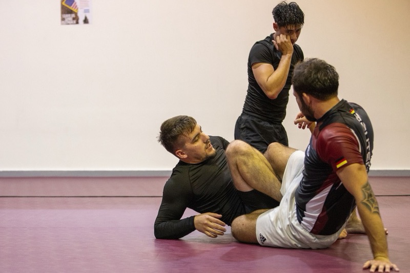 An instructor sits on the mat explaining a seated guard position to two students.