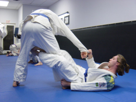 The bottom player hooks the standing opponent's lead leg from the outside in De La Riva guard.
