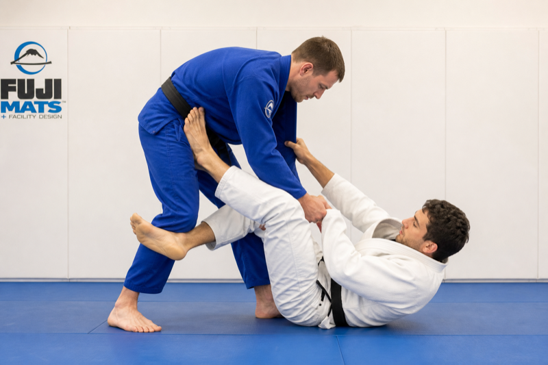 The bottom player wraps the standing opponent's near ankle from the inside in reverse De La Riva.