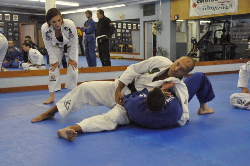 A grappler in a white gi pins a partner from side control while an instructor watches.