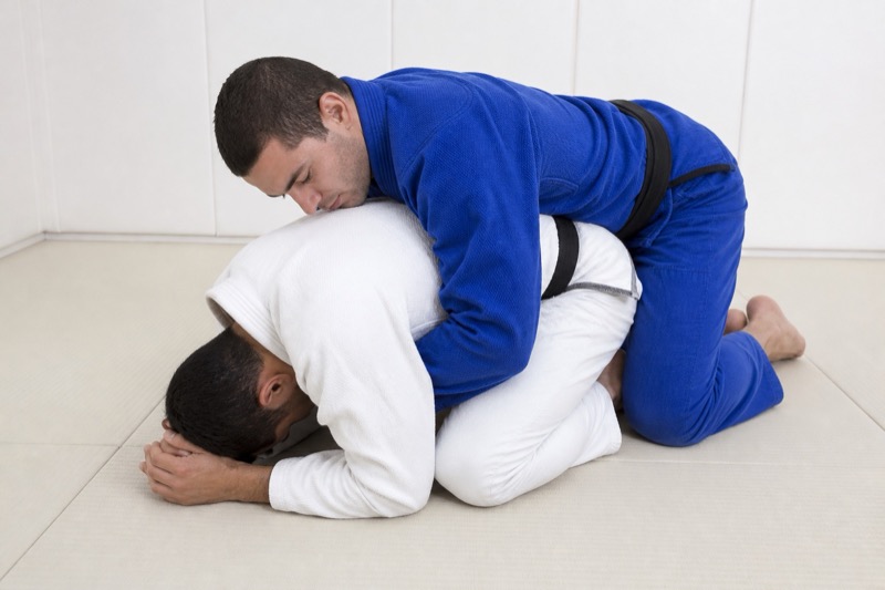 One grappler is on hands and knees in turtle while the other drapes a seatbelt over their back.