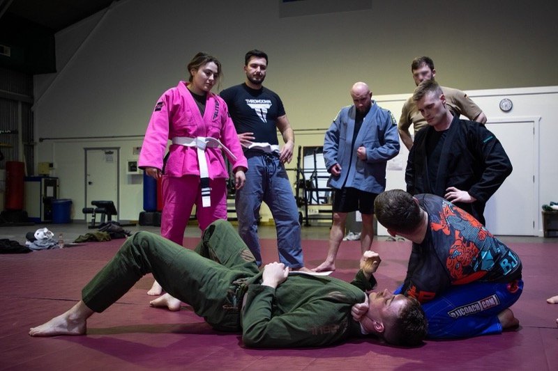 Students in mixed gis watch an instructor demonstrate a technique on the mat.